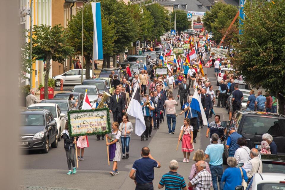 Eine lebhafte Straßenparade, bei der die Teilnehmer Transparente und Fahnen tragen und zwischen geparkten Autos hindurchziehen – das ist das Wiesenfest in Weißenstadt. Die Zuschauer säumen die Bürgersteige, und in der von Bäumen gesäumten Straße herrscht eine festliche und gesellige Atmosphäre.