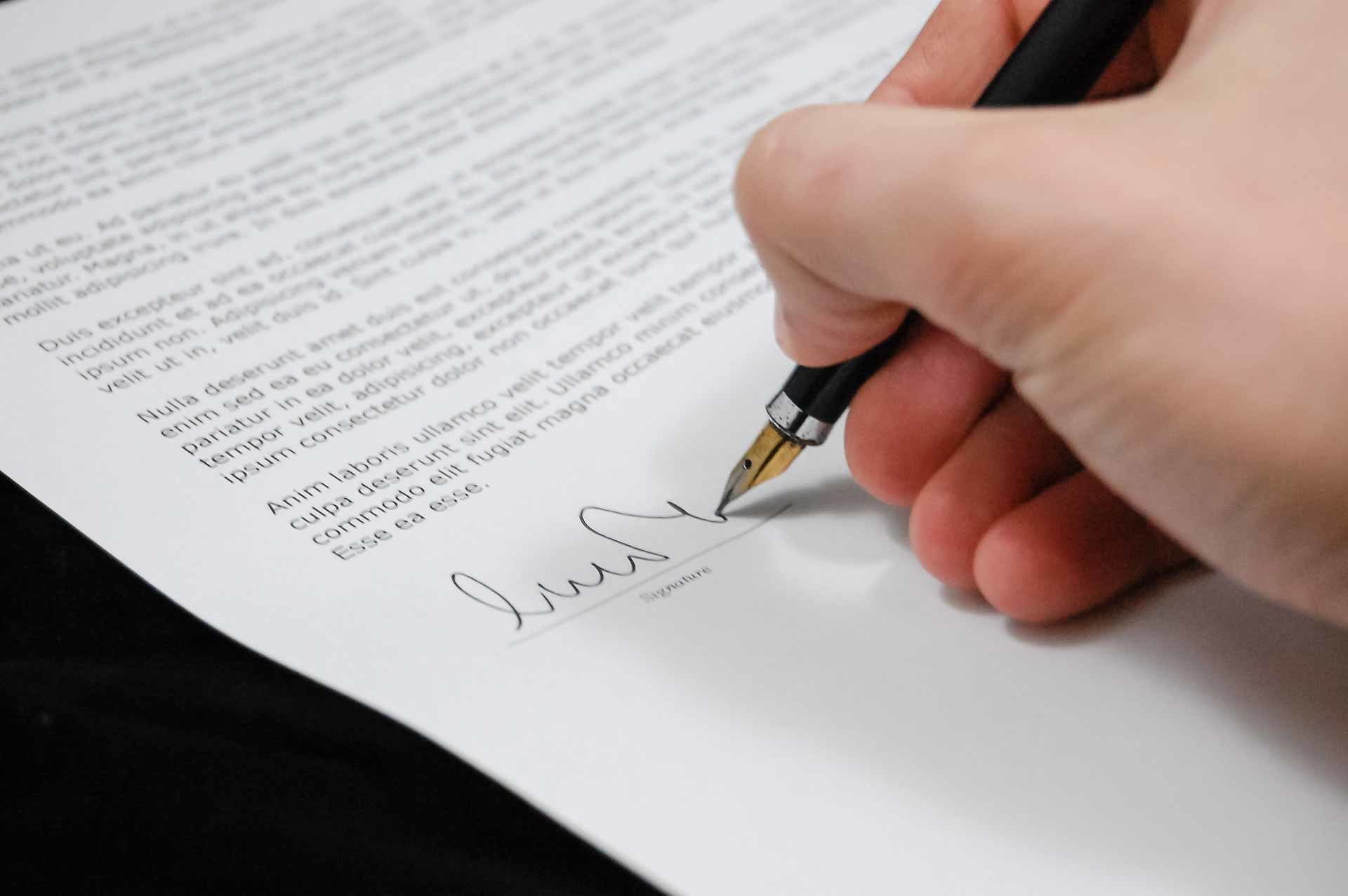 Eine Hand, die einen Füllfederhalter hält, unterschreibt ein gedrucktes Dokument auf weißem Papier, möglicherweise eine Datenschutzerklärung. Nur die Unterschriftszeile, der Stift und ein Teil der Hand der Person sind scharf abgebildet.
