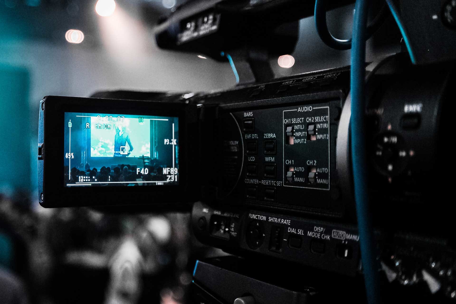 Nahaufnahme einer professionellen Videokamera, die eine Veranstaltung für „Medien Bad Weißenstadt am See“ aufzeichnet; auf dem Sucherbildschirm der Kamera sind die Bühne und das Publikum scharf abgebildet, der Hintergrund ist unscharf.