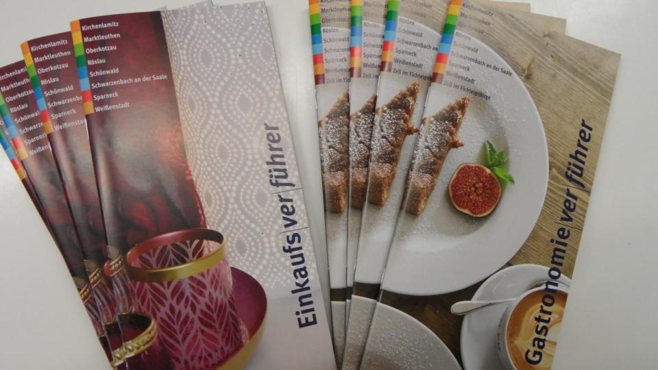 Mehrere bunte deutsche Broschüren; auf dem Umschlag des einen Stapels sind Teetassen abgebildet, auf dem anderen ein Teller mit Dessert und Feigen; auf beiden ist deutscher Text zu erkennen.