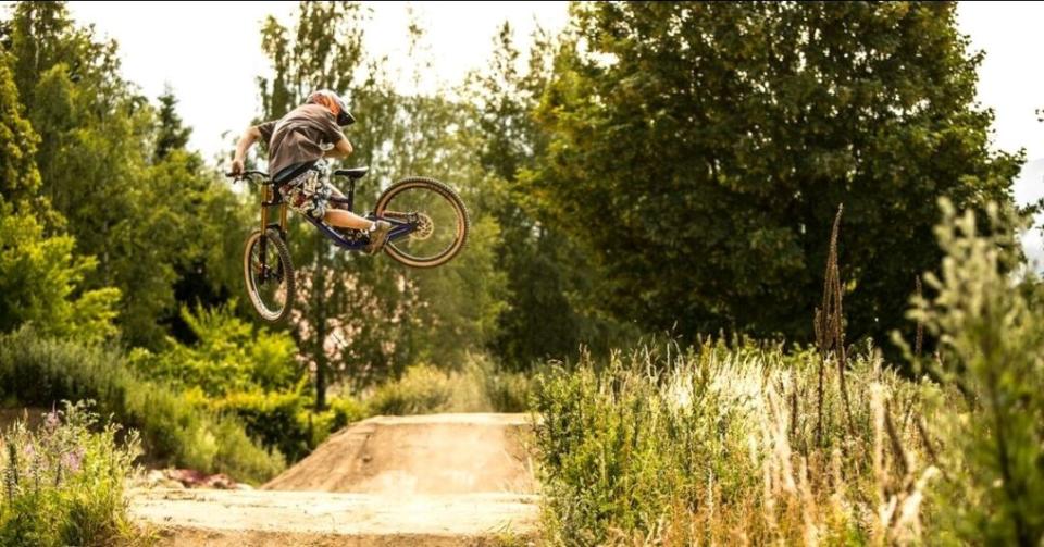 Eine Person mit Helm führt auf einem Mountainbike einen Sprung über einen Feldweg aus, umgeben von hohem Gras, Bäumen und strahlendem Sonnenschein.