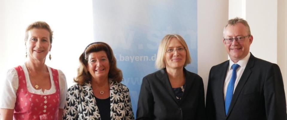 Vier Erwachsene – drei Frauen und ein Mann – stehen vor einem blau-weißen Heilwald-Hintergrund und lächeln in die Kamera. Die Frauen tragen festliche Kleidung, eine davon in traditioneller Tracht, und der Mann trägt Anzug und Krawatte.