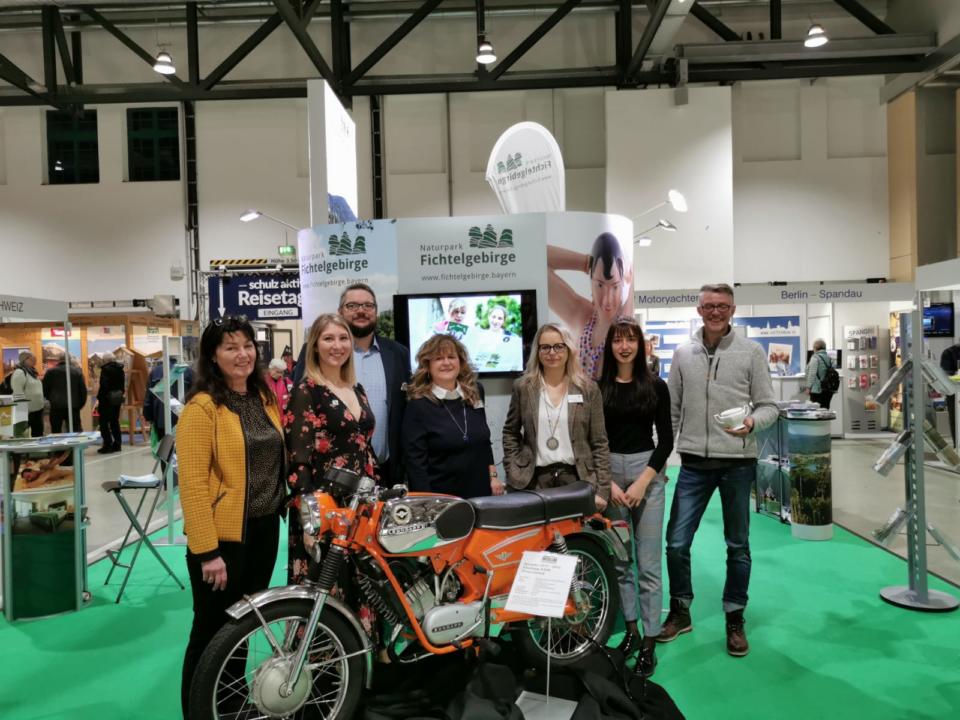 An einem Messestand in der Halle in Weißenstadt im Rahmen der Reisemesse Dresden stehen sieben Menschen lächelnd um ein orangefarbenes Oldtimer-Motorrad herum, umgeben von Informationsbannern und einem grünen Teppich.
