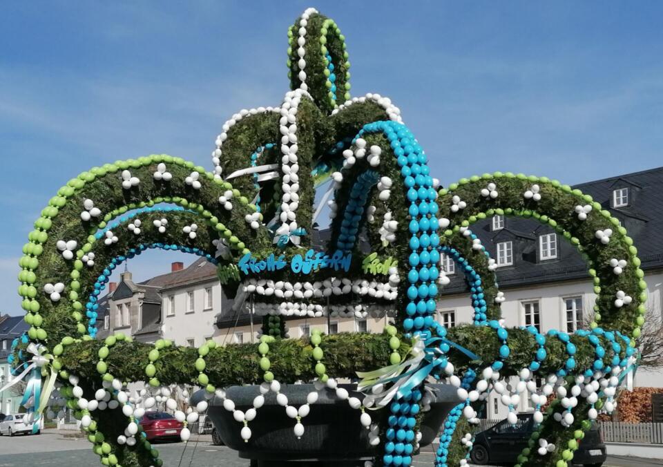 Ein großer Osterbrunnen im Freien ist mit grünen Girlanden und weißen, blauen und grünen Ostereiern geschmückt, die unter strahlend blauem Himmel in Bögen angeordnet sind.