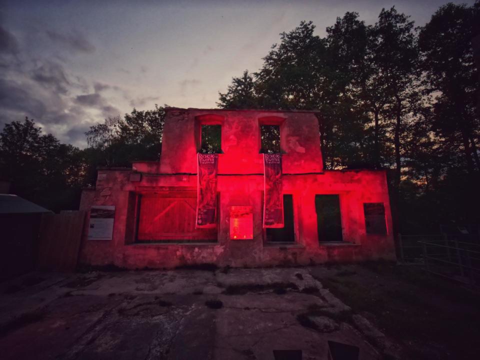 Ruinen eines Gebäudes, das in der Abenddämmerung von unheimlichen roten Lichtern beleuchtet wird, mit Bäumen und einem bewölkten Himmel im Hintergrund. Die Fenster und Türen sind leer, was dem Bauwerk ein gespenstisches Aussehen verleiht, als wäre es während einer „Night of Light“-Veranstaltung fotografiert worden.