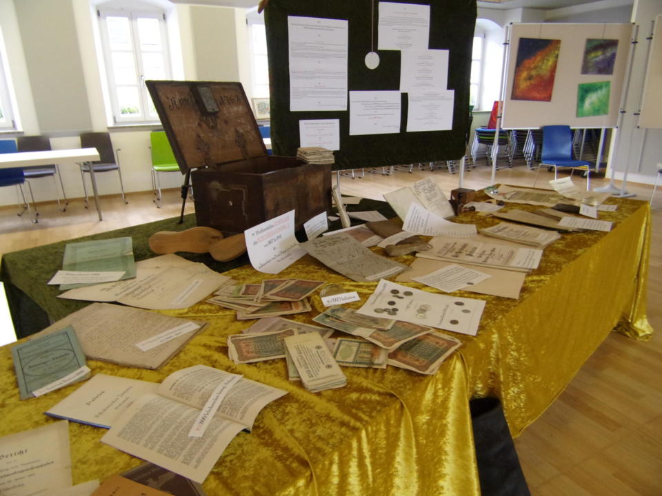 Auf einem mit einem goldenen Tuch bedeckten Ausstellungs-Tisch liegen alte Papiere, Dokumente, Bücher, Münzen und Banknoten. Dahinter sind eine Truhe und eine Tafel mit daran befestigten Papieren zu sehen, im Hintergrund zieren farbenfrohe Kunstwerke die Wände.