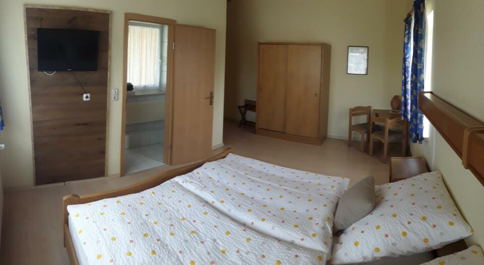 Ein ordentlich gemachtes Bett mit gepunkteter Bettwäsche in einem schlichten Schlafzimmer im Landgasthaus Birkenhof, ausgestattet mit Holzmöbeln, einem an der Wand montierten Fernseher, einem Kleiderschrank, einem Schreibtisch, einem Stuhl und einer Tür, die zu einem gefliesten Bereich führt.