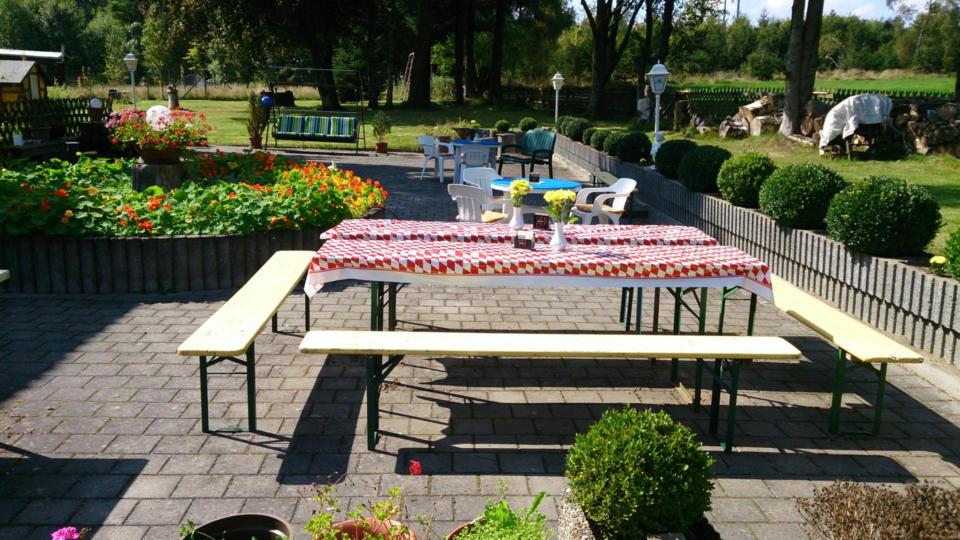 In einer sonnigen Gartenterrasse, umgeben von Blumen, gepflegten Sträuchern und weiteren Sitzgelegenheiten, stehen zwei lange Bänke und ein Tisch mit einer rot karierten Tischdecke. Im Hintergrund sind Bäume und Rasenflächen zu sehen.