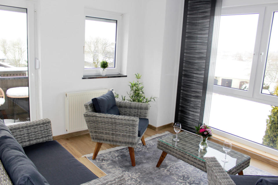 Modernes Wohnzimmer mit Korbmöbeln, blauen Kissen, einem Couchtisch aus Glas, Blumen, Weingläsern, Topfpflanzen, großen Fenstern und einem Blick vom Balkon auf die verschneite Landschaft. Neutrale Farben und natürliches Licht prägen den Raum.