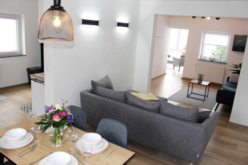 Moderner, offener Wohn- und Essbereich mit einem grauen Sofa, einem Holztisch für vier Personen, frischen Blumen und hellem Tageslicht, das durch große Fenster hereinströmt.