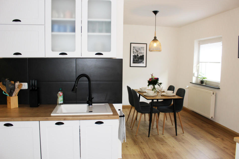 Moderne Küche mit weißen Schränken, schwarzer Armatur, Küchenutensilien und Olivenöl neben der Spüle. Im Hintergrund steht ein Esstisch, der mit Geschirr und Blumen gedeckt ist; darüber hängt eine Lampe. Auf der Fensterbank steht eine Pflanze.