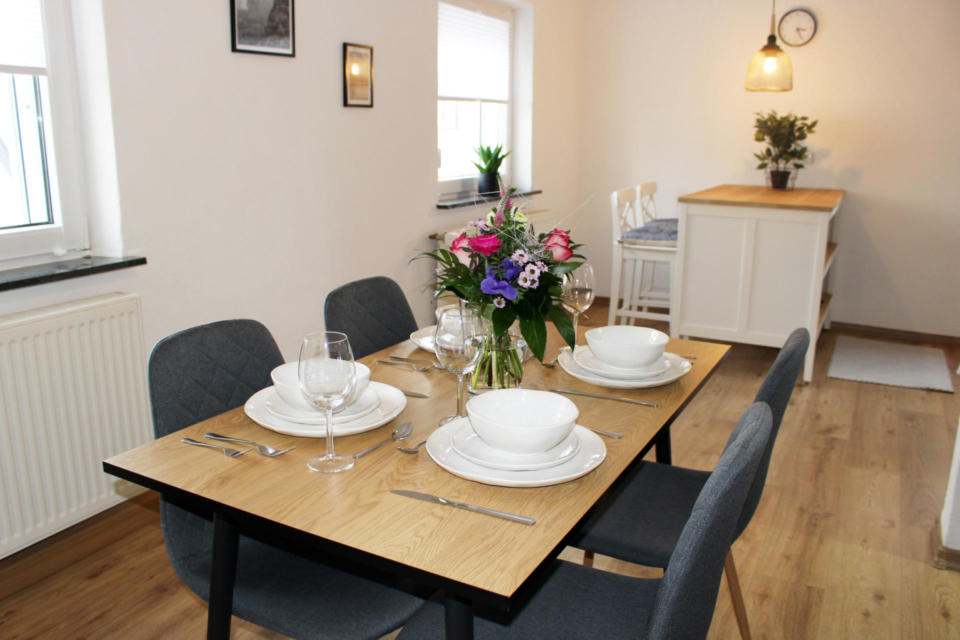 Ein modernes Esszimmer-Set für vier Personen mit weißem Geschirr, Weingläsern, Besteck und einer Vase mit bunten Blumen. Graue Stühle stehen um den Tisch herum in einem hellen Raum mit Holzboden und minimalistischer Einrichtung.