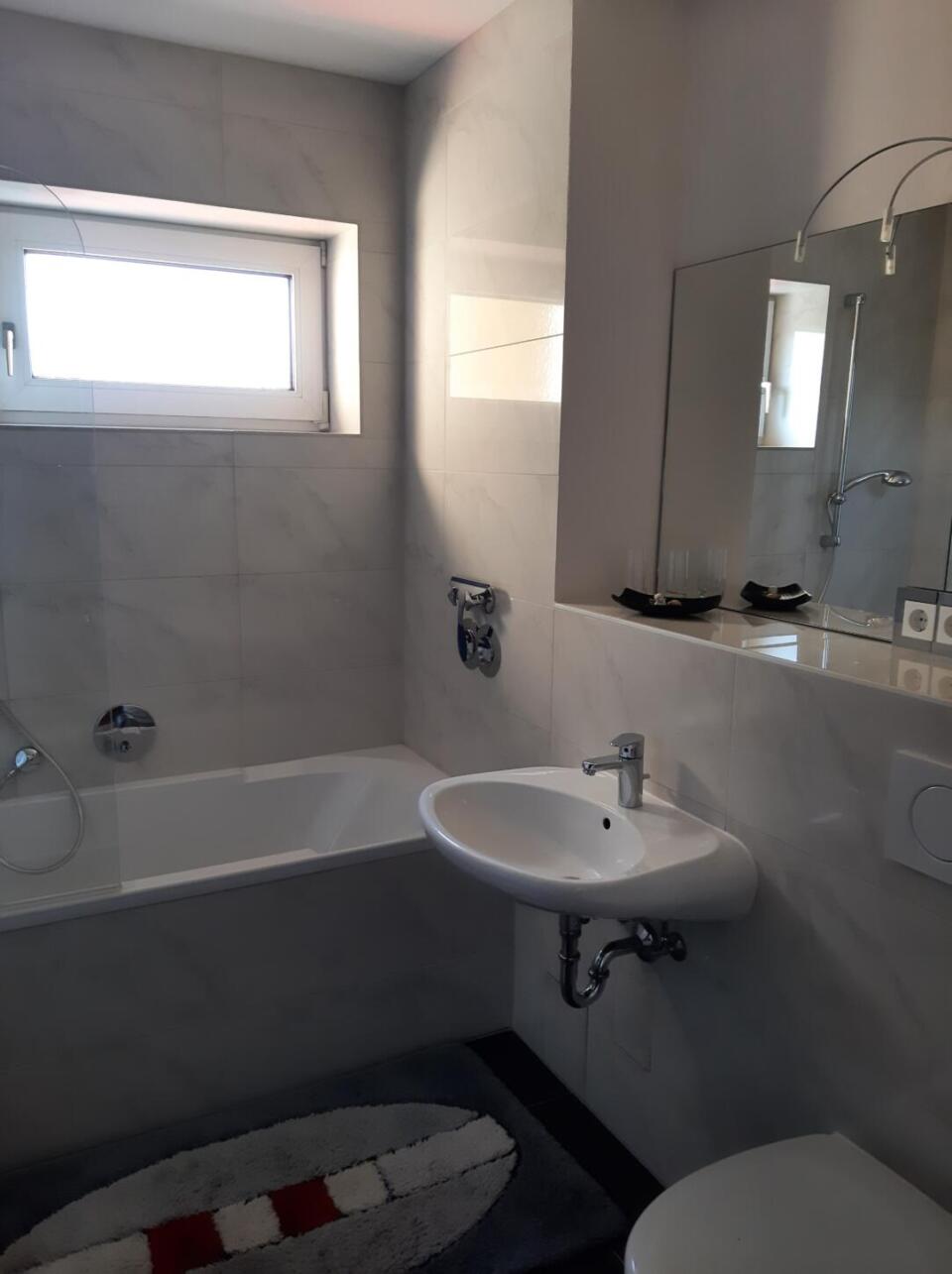 Modernes Badezimmer mit Badewanne, wandmontiertem Waschbecken, Spiegel und einem kleinen Fenster in der stilvollen Ferienwohnung „Sunset“. Auf dem Boden liegt ein grauer Teppich mit roten und weißen Streifen, der zusammen mit den hell gefliesten Wänden eine helle und einladende Atmosphäre schafft.
