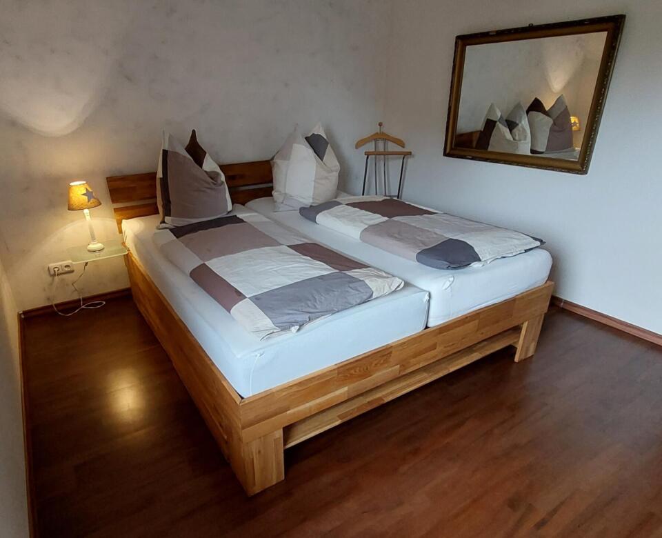 Ein aufgeräumtes Schlafzimmer in der Ferienwohnung „Sunset“ verfügt über zwei aneinandergeschobene Einzelbetten, braune und weiße Bettwäsche, Kissen, einen kleinen Nachttisch mit einer Lampe, einen großen Wandspiegel und Holzböden.