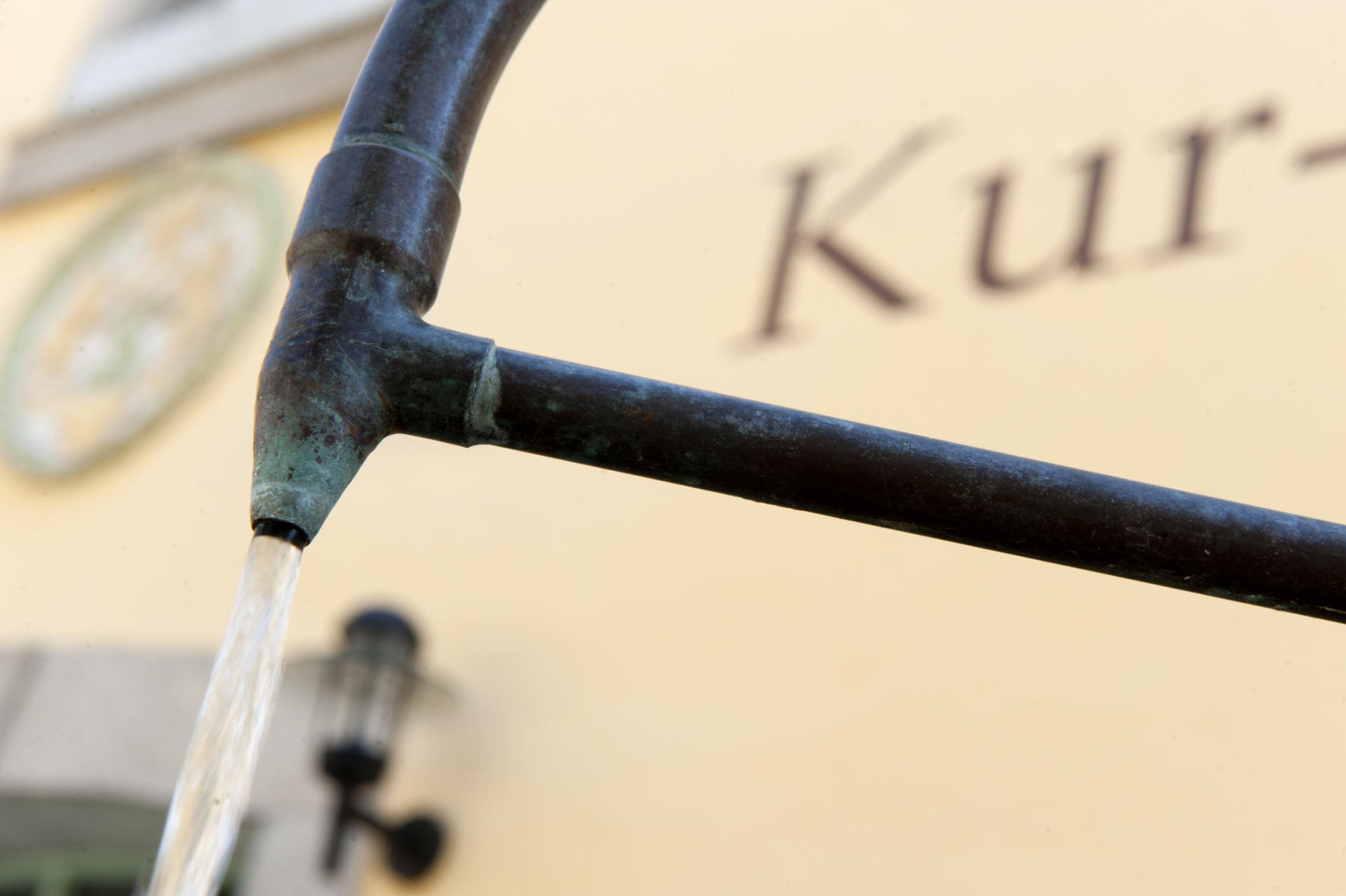 Eine Nahaufnahme von Wasser, das aus einem verwitterten Metallrohr fließt, mit einer unscharfen Gebäudefassade, einer Lampe, einem runden Schild und dem teilweise sichtbaren Schriftzug „Kur-“ im Hintergrund – ein ruhiger Moment, wie man ihn bei einer Führung am Weißenstädter See erleben könnte.