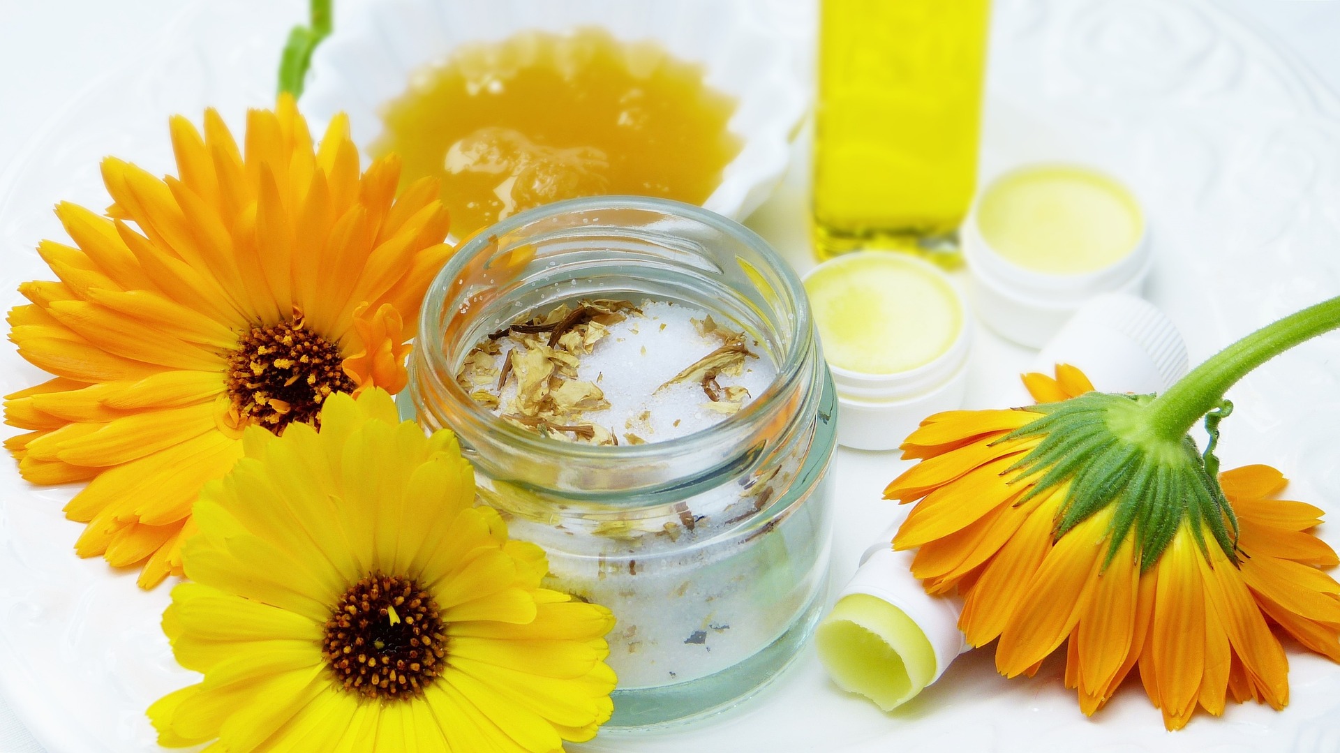 Eine Nahaufnahme von gelben und orangefarbenen Ringelblumen, ein Glas mit Kräutercreme, Honig, eine kleine Flasche Öl und Lippenbalsam-Tiegel, angeordnet auf einer weißen Unterlage – die perfekte Inspiration für einen Workshop für Kinder und Jugendliche zum Thema natürliche Hautpflege und Wellness.