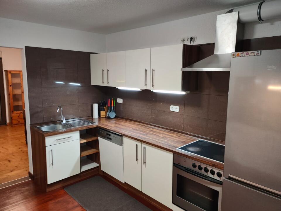 Moderne Küche mit weißen Schränken, braunen Arbeitsplatten, Edelstahlgeräten, Spüle, Elektroherd, Dunstabzugshaube und Kühlschrank. Eine Unterbauleuchte beleuchtet den Arbeitsbereich. Im angrenzenden Raum ist ein Holzboden zu sehen.