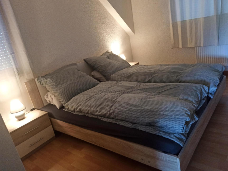 Ein ordentlich gemachtes Bett mit gestreifter Bettwäsche, zwei Kissen auf jeder Seite und Nachttischen mit brennenden Lampen auf beiden Seiten. Das Zimmer hat helle Wände, Holzböden und ein Fenster mit transparenten Vorhängen.