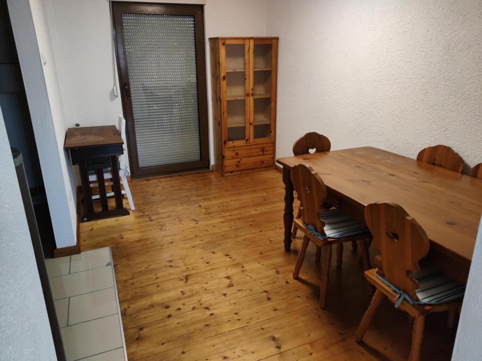 Ein kleines Esszimmer mit einem Holztisch, vier Stühlen mit gestreiften Kissen, einer Vitrine mit Glasfront, einem Beistelltisch, Holzboden und einem großen Fenster mit geschlossenen Jalousien.