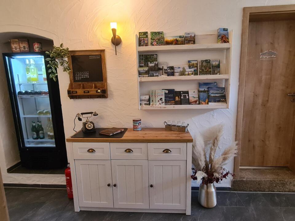 Gemütlicher Eingangsbereich mit einem weißen Schrank, einem Vintage-Telefon und Broschüren auf Wandregalen. Daneben steht ein Kühlschrank mit Glasfront, in dem Getränke aufbewahrt werden, und auf dem Boden steht eine Vase mit Trockenblumen. Warme Beleuchtung sorgt für eine einladende Atmosphäre.