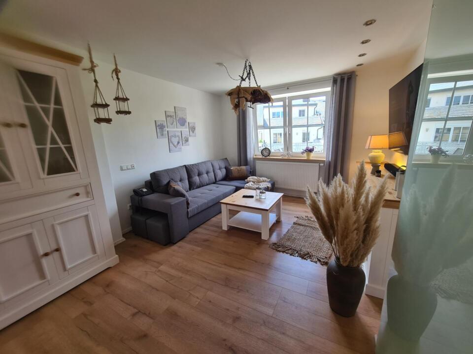 Helles Wohnzimmer mit grauem Sofa, weißem Couchtisch, Pampasgras in einer Vase, Holzboden, Wanddekoration, einem weißen Schrank und einem Fenster mit grauen Vorhängen, durch das Tageslicht hereinströmt.