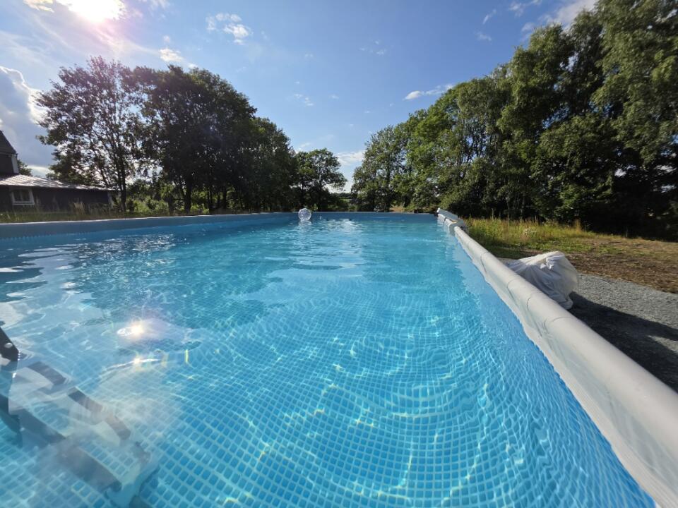 Im Freien befindet sich ein Aufstellpool, gefüllt mit klarem, blauem Wasser, umgeben von Bäumen und Rasen, auf dessen Oberfläche sich das Sonnenlicht spiegelt.