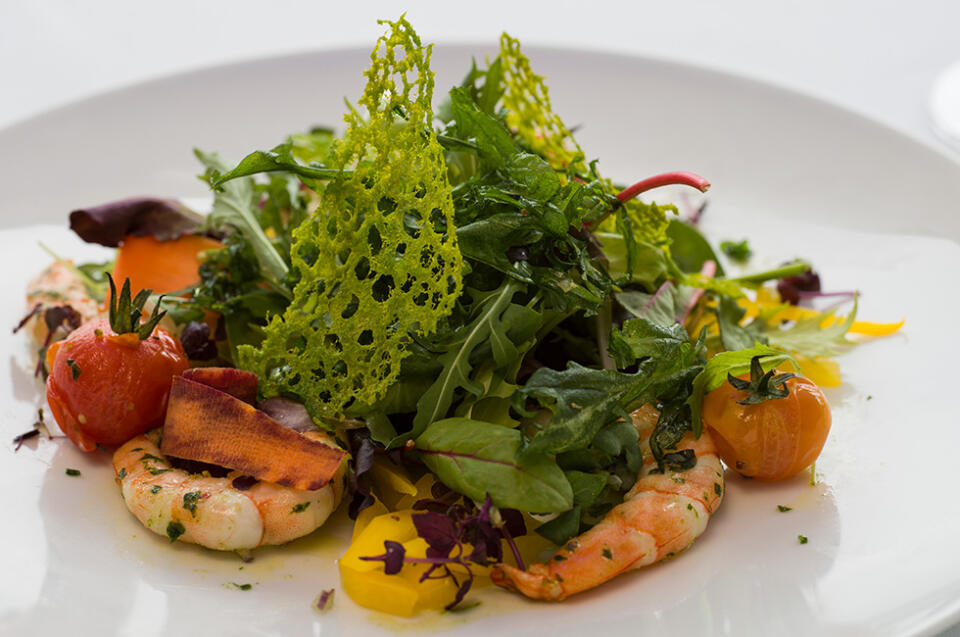Ein Gourmet-Salat mit Garnelen, gemischtem Blattsalat, gelben und roten Kirschtomaten, Karottenscheiben und einer knackigen grünen Beilage wird im SeeBlick Siebenquell elegant auf einem weißen Teller angerichtet.