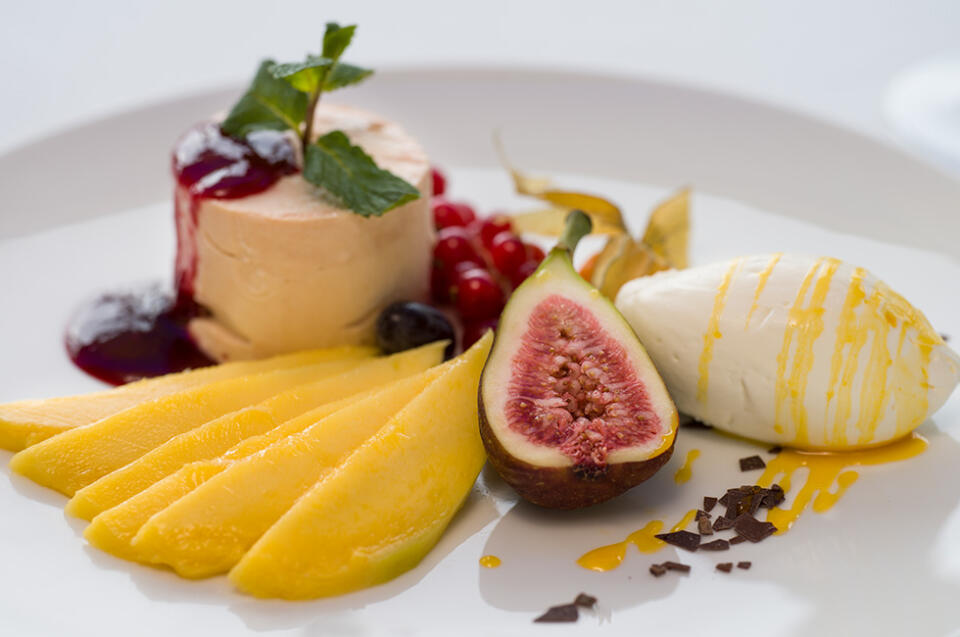 Ein Dessertteller im SeeBlick Siebenquell besteht aus Mangoscheiben, einer halbierten Feige, Schlagsahne mit gelbem Sirup, roten Johannisbeeren und einem runden Kuchen, der mit Beerensauce und Minzblättern garniert ist.