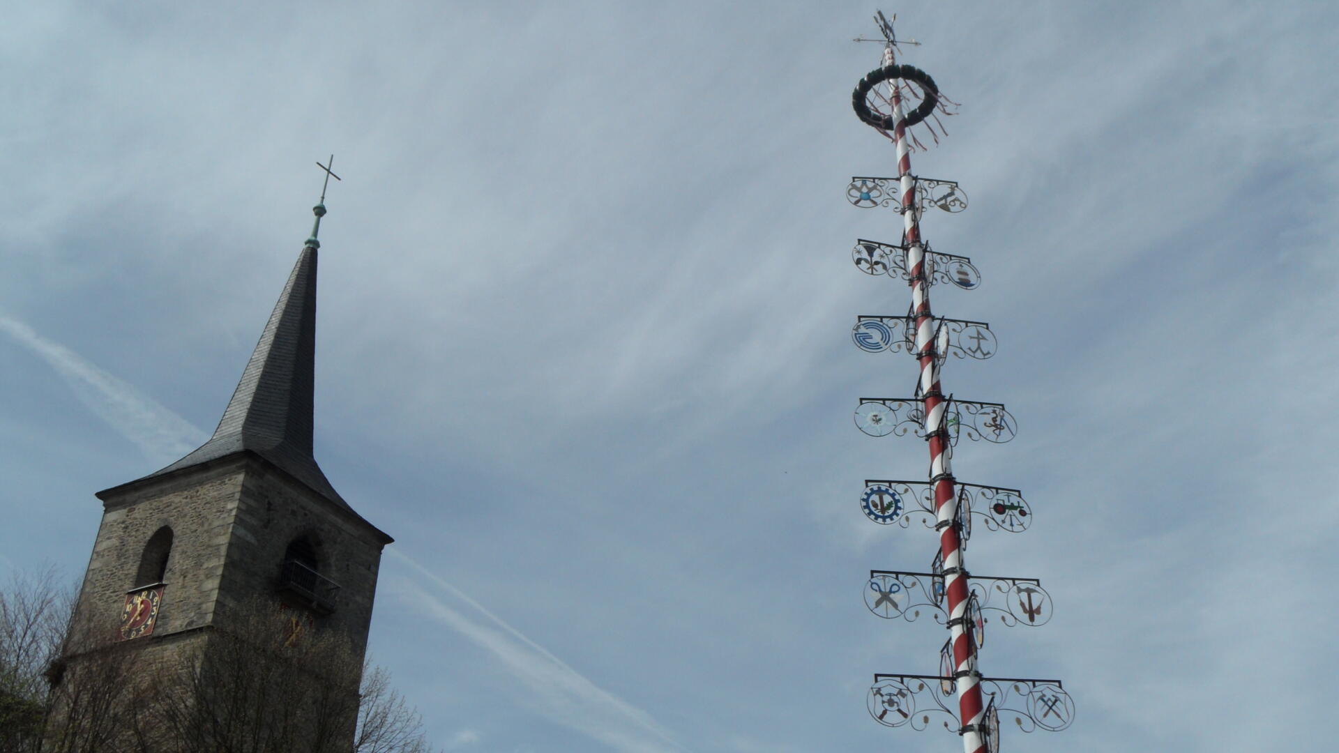 Ein hoher, mit bunten Symbolen geschmückter Maibaum steht neben einem steinernen Kirchturm mit einem Kreuz an der Spitze – beide vor einem bewölkten Himmel –, was die festliche Stimmung des Maibaumfestes in Weißenstadt einfängt.