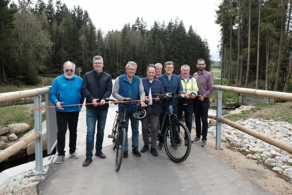 Acht Personen stehen auf einer neuen Brücke entlang des Radwegs, einige halten Fahrräder in der Hand, und gemeinsam durchschneiden sie ein orangefarbenes Band, um die offizielle Eröffnung des Egerradwegs zu feiern. Im Hintergrund sind Wald und Felsen zu sehen.