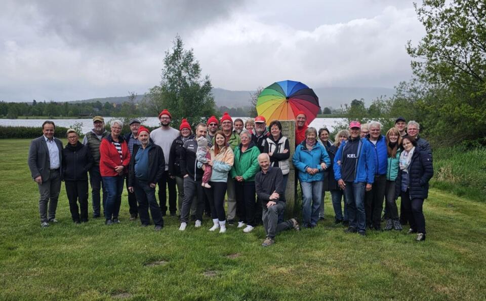 Eine Gruppe von etwa 30 Personen steht auf einer Wiese und lächelt in die Kamera. Die meisten tragen Jacken, einige haben rote Mützen auf und gehören zu den Motorradfreunden Wunsiedel. In der Mitte steht ein bunter Regenschirm. Im Hintergrund sind unter einem bewölkten Himmel Bäume und ein See zu sehen.