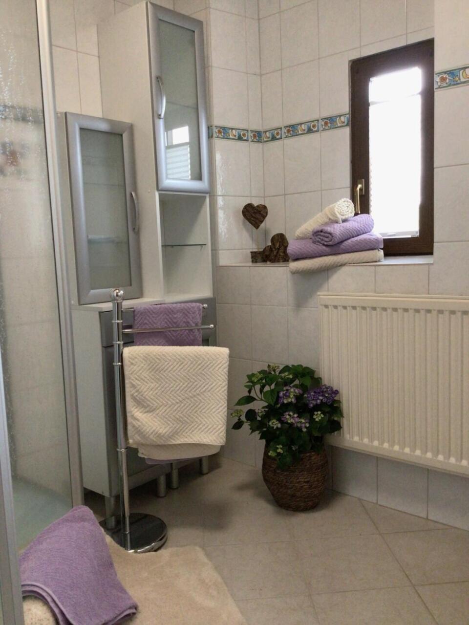 Ein aufgeräumtes Badezimmer mit weißen Fliesen, gefalteten lila-weißen Handtüchern, einem hohen Schrank, einem Fenster, einem Heizkörper und einer Topfpflanze mit lila Blüten auf dem Boden. In der Nähe hängen weitere Handtücher an einem Handtuchhalter.