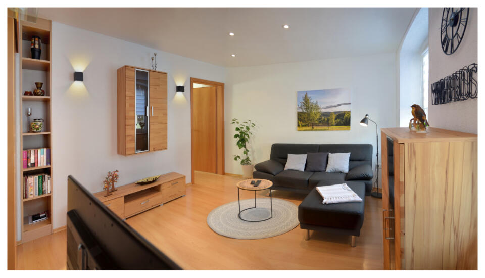 Modernes Wohnzimmer mit hellen Holzböden, grauem Sofa, kleinem rundem Teppich, Wandregalen, Bücherregal, Fernseher, Topfpflanze, Wandbildern und Dekoartikeln. Neutrale Farbtöne und helle Beleuchtung schaffen eine gemütliche Atmosphäre in dieser charmanten Ferienwohnung in Fuss.