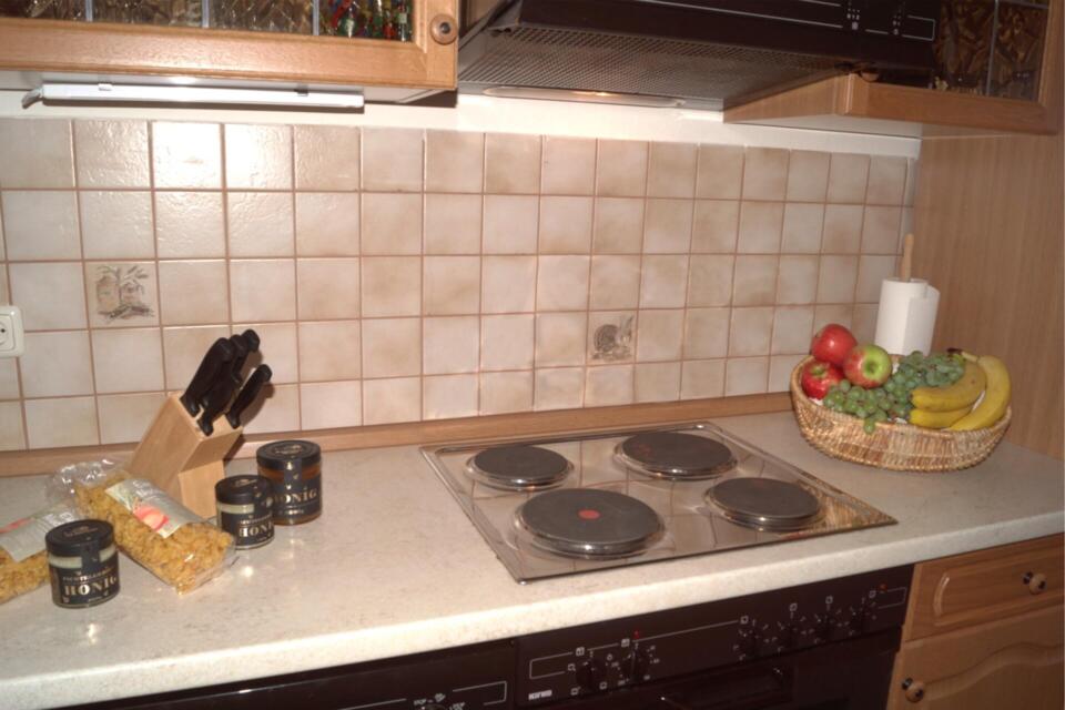 Eine Küchenarbeitsplatte mit einem Elektroherd, einem Messerblock, Gläsern, Nudeln, einem Küchenrollenhalter und einem Obstkorb voller Äpfel, Trauben und Bananen vor einer gefliesten Küchenrückwand.