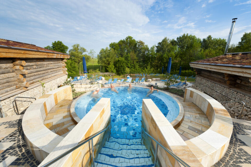 Außen-Whirlpool in der 5Kurfranken Therme, in dem sich Menschen im warmen, blauen Wasser entspannen, umgeben von Stein- und Holzkonstruktionen, grünen Bäumen und einem blauen, mit Wolken übersäten Himmel. Vom Vordergrund aus führen Stufen hinunter in das runde Becken.