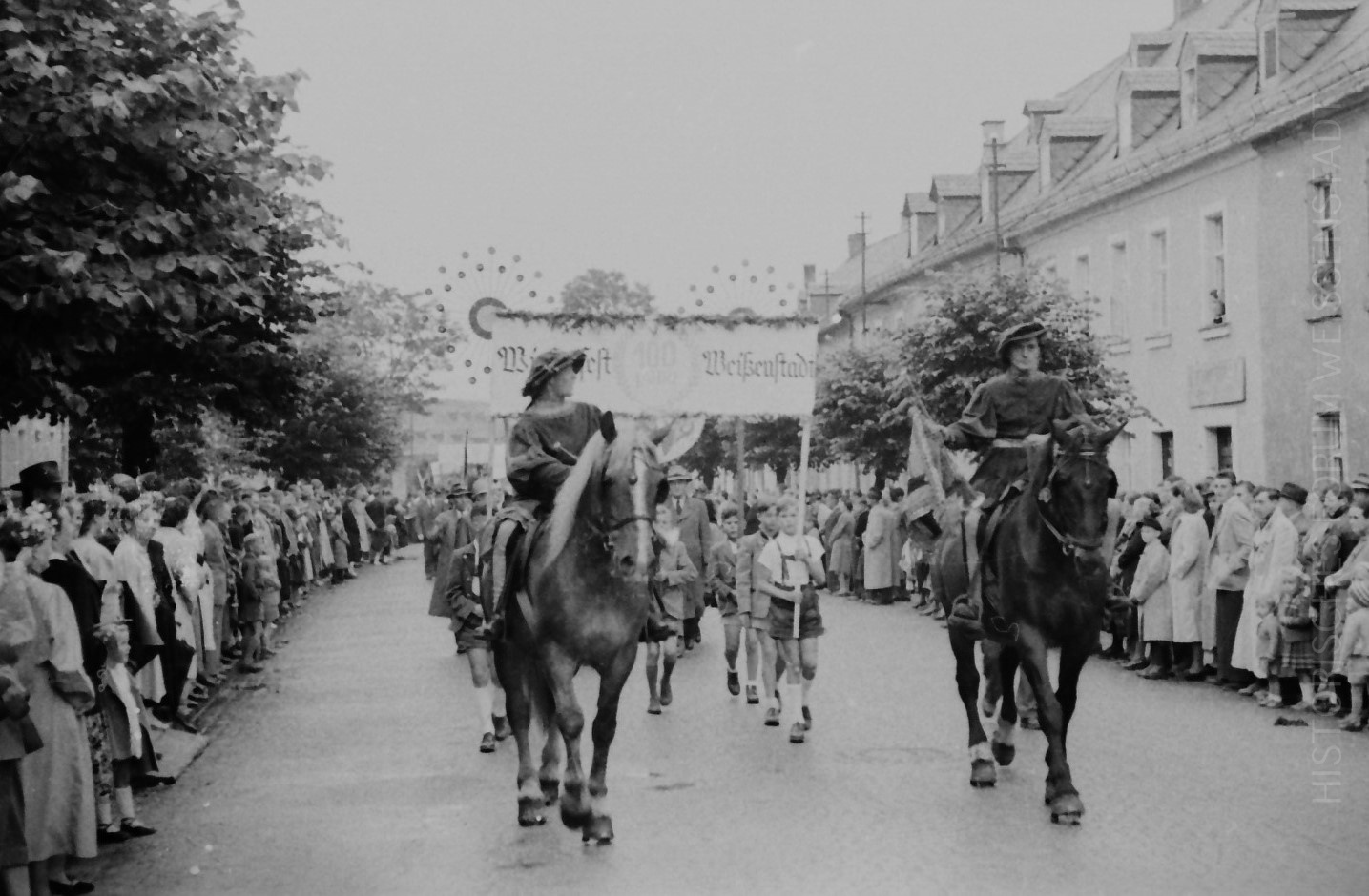 Schwarz-Weiß-Foto einer Straßenparade, bei der zwei Personen zu Pferd eine Gruppe anführen, während auf beiden Seiten Menschenmengen zuschauen; im Hintergrund ist auf der anderen Straßenseite ein Transparent zu sehen, das die lebhafte Stimmung einer Veranstaltung im Historischen Forum Weißenstadt einfängt.