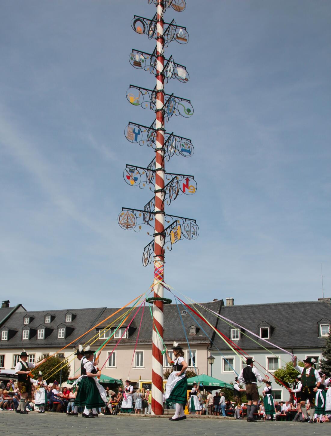 Menschen in traditioneller Tracht tanzen auf einem Stadtplatz um einen mit bunten Bändern geschmückten Maibaum. Im Hintergrund sind Gebäude und ein klarer Himmel zu sehen, und am hohen Maibaum hängen verzierte Schilder.