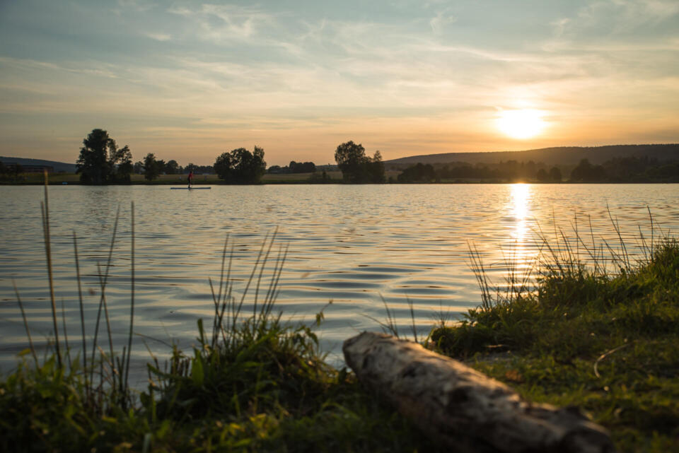 Ein ruhiger See bei Sonnenuntergang mit sanften Wellen auf der Wasseroberfläche bildet eine friedliche Kulisse zum Nachdenken, die an die Gelassenheit erinnert, die die „Bürgerbefragung Titel Weißenstadt“ hervorruft. Bäume säumen das gegenüberliegende Ufer, und in der Ferne ist eine Person beim Stand-up-Paddling zu sehen, während im Vordergrund Gras und ein Baumstamm zu erkennen sind, die diese friedliche Landschaft vervollständigen.