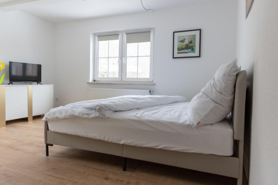 Ein minimalistisches Schlafzimmer mit einem ordentlich gemachten Bett, weißer Bettwäsche, einem Fernseher auf einem weißen Ständer, Holzboden, einem Fenster, durch das Tageslicht hereinströmt, und einem gerahmten Kunstwerk an der Wand – ganz im Zeichen der ruhigen Eleganz des Wulgerahof-Stils.