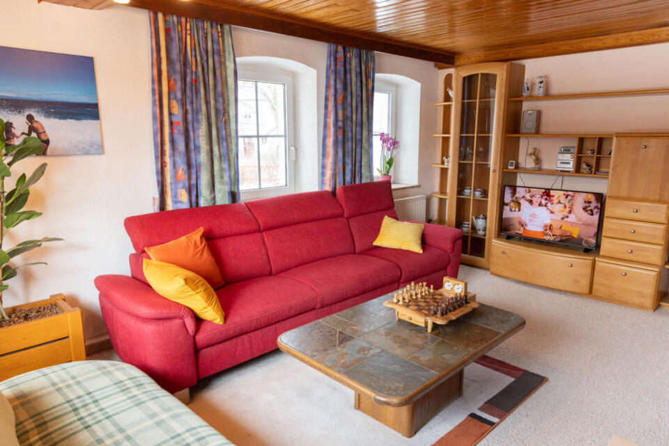 Das gemütliche Wohnzimmer im Wulgerahof ist mit einem roten Sofa, Holzregalen, einem Couchtisch mit einem Schachspiel, einem Fernseher, auf dem eine Sendung läuft, Topfpflanzen, bunten Vorhängen und einem Strandfoto an der Wand ausgestattet. Durch zwei Fenster fällt Sonnenlicht herein.