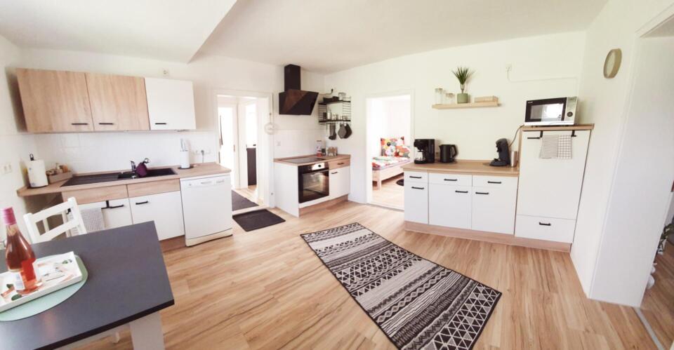 Moderne Küche mit Holzböden, weißen Schränken, einem gemusterten Teppich, Backofen, Spüle, Einbaugeräten und einer Mikrowelle. In der Ecke steht ein Esstisch mit einem Tablett. Die helle Atmosphäre dieser Ferienwohnung am See setzt sich im angrenzenden Schlafzimmer fort, wo bunte Bettwäsche für eine einladende Note sorgt, die durch die offene Tür zu sehen ist.