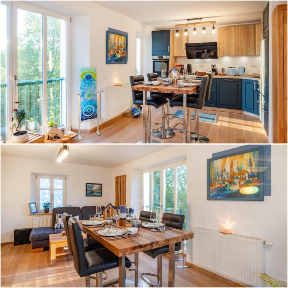 Die moderne Ferienwohnung „Ferienhaus Die Blaue Pause“ verfügt über Holzböden, eine Küche in Blau- und Holztönen, einen Esstisch für vier Personen, helle Fenster, Kunstwerke an den Wänden sowie einen gemütlichen Wohnbereich mit einem Sofa und kleinen Dekoartikeln.