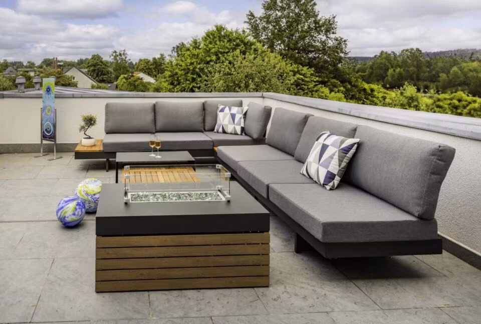 Moderne Außenterrasse in der Ferienwohnung Die Blaue Pause 42 mit einem grauen Sektionssofa, geometrischen Kissen, einem Tisch mit Feuerstelle, zwei Weingläsern und bunten Dekokugeln, alles umgeben von Grünpflanzen unter einem bewölkten Himmel.
