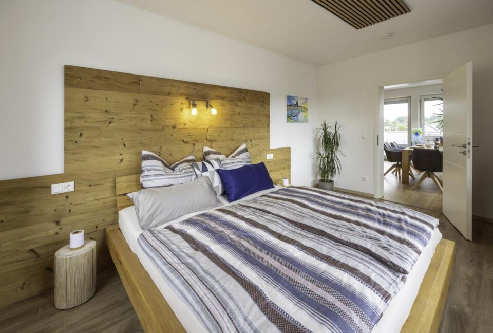 Modernes Schlafzimmer in der Ferienwohnung Die Blaue Pause 42 mit einem großen Bett mit gestreiftem Bettzeug, einem Kopfteil aus Holz und kleinen Nachttischen. Der helle Raum öffnet sich zu einem angrenzenden Raum mit Pflanzen und einem schönen Blick nach draußen.