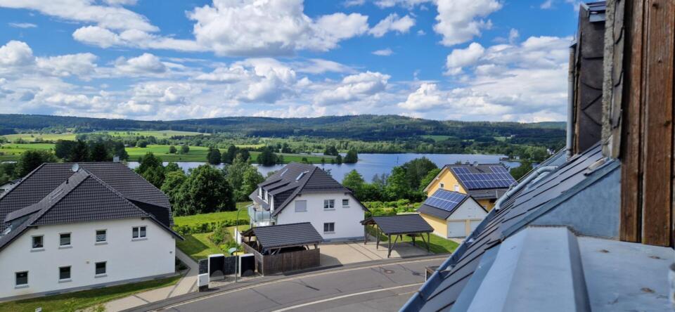 Ein malerischer Ausblick auf Häuser mit dunklen Dächern, eine kurvenreiche Straße, einen See, grüne Felder und in der Ferne bewaldete Hügel unter einem blauen Himmel mit vereinzelten Wolken – das ist die idyllische Kulisse rund um die Ferienwohnung „überm See Weißenstadt“.