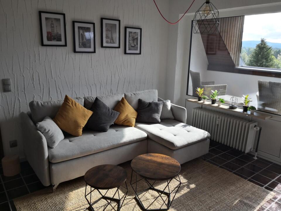 Das gemütliche Wohnzimmer der Ferienwohnung „überm See“ in Weißenstadt verfügt über ein graues Ecksofa, gelbe und graue Kissen, runde Holztische, stilvolle Wandbilder, ein großes Fenster mit Blick auf Bäume sowie Pflanzen auf der Fensterbank.
