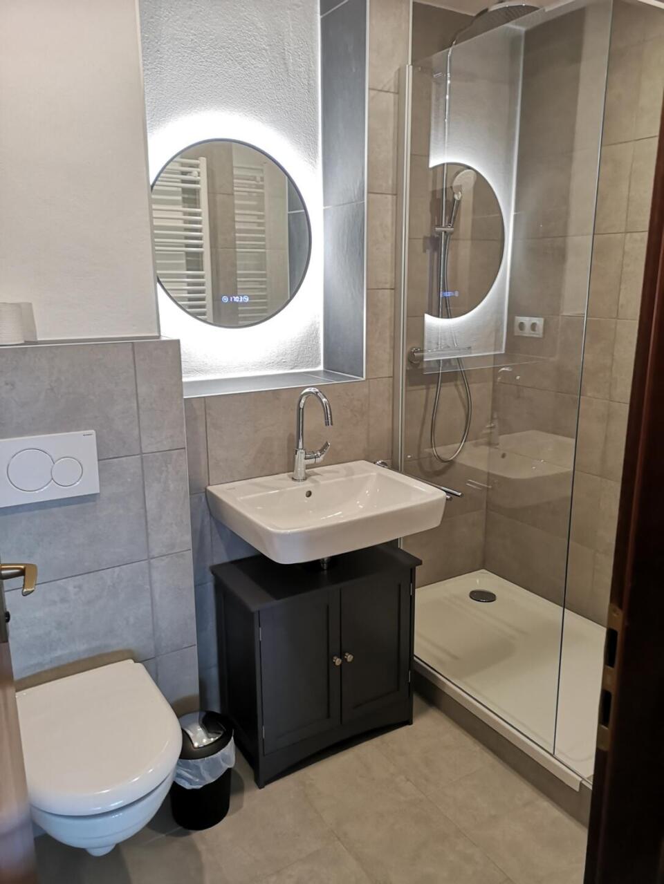 Modernes Badezimmer in der Ferienwohnung „Überm See“ in Weißenstadt, ausgestattet mit einem wandhängenden WC, einem quadratischen Waschbecken auf einem dunklen Unterschrank, einem runden Spiegel mit LED-Beleuchtung und einer Duschkabine aus Glas mit Handbrause.