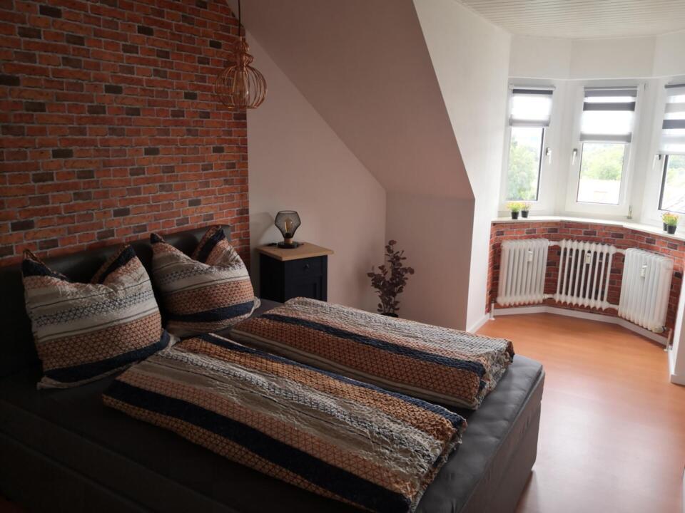 Modernes Schlafzimmer in der Ferienwohnung „Überm See“ in Weißenstadt, mit einer auffälligen Akzentwand aus Ziegelsteinen, einem Doppelbett mit gemusterter Bettwäsche, einem kleinen schwarzen Nachttisch, einer dekorativen Lampe und einem Erkerfenster, das mit weißen Jalousien und Topfpflanzen auf der Fensterbank geschmückt ist.