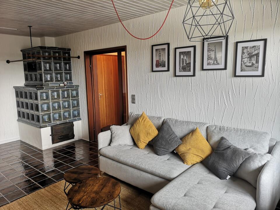 Das gemütliche Wohnzimmer der Ferienwohnung „überm See Weißenstadt“ ist mit einem hellgrauen Sofa, gelben und grauen Kissen, einem runden Couchtisch aus Holz und einem gekachelten Holzofen in der Ecke ausgestattet. Gerahmte Kunstwerke schmücken die weißen, strukturierten Wände, ergänzt durch moderne Pendelleuchten, die von der Decke hängen.
