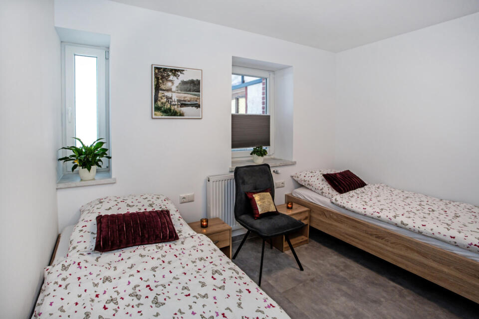 Ein helles Schlafzimmer mit zwei Einzelbetten, jeweils mit geblümter Bettwäsche und einem burgunderroten Kissen, die durch einen schwarzen Stuhl voneinander getrennt sind. Eine Pflanze, ein gerahmtes Bild und Kerzen schmücken den Raum. Es gibt zwei Fenster und weiße Wände.